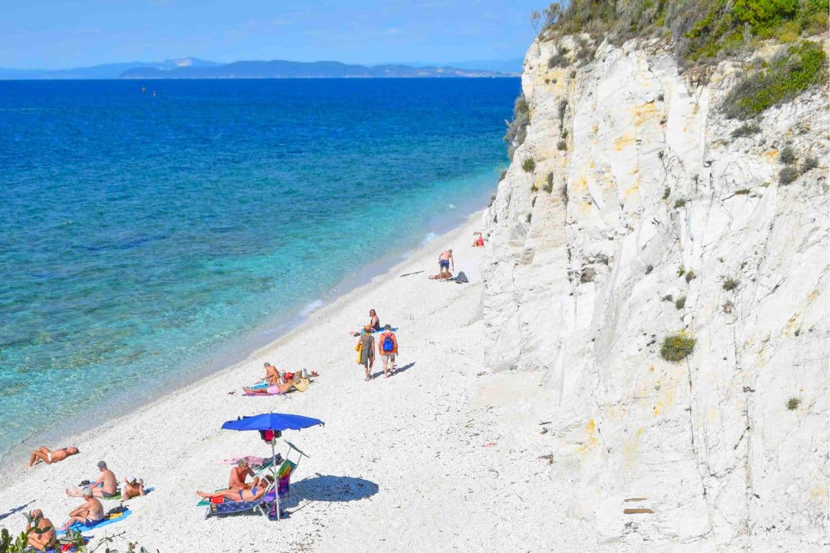 People sunbathing on a bright white sandy beach framed by a white cliff and turquoise sea on Elba Island Italy