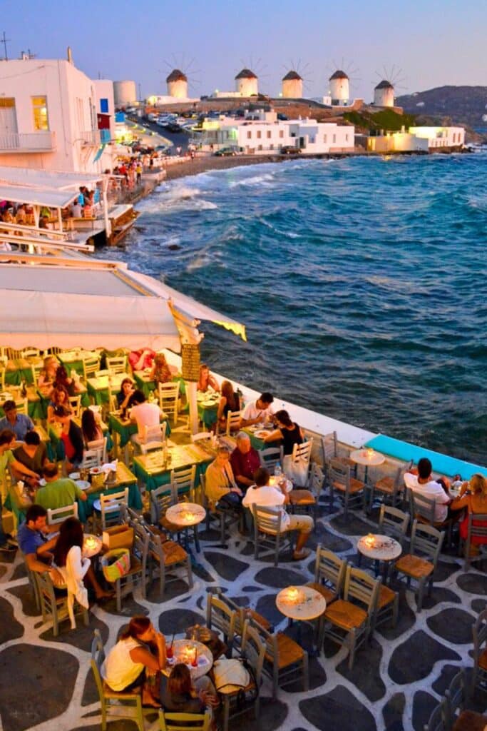 Image of people sitting at cafe tables on the water at dusk with the windmills of Mykonos Greece in the background