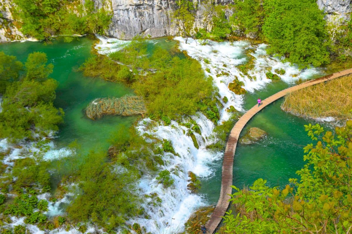 A woman in a pink jacket walks a winding wooden path across a lake surrounded by waterfalls at Plitvice Lakes National Park