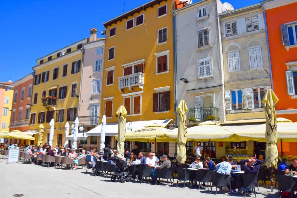 People sit a sunny cafe tables in front of colorful buildings in the town of Rovinj
