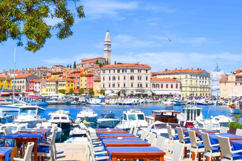 Colorful cafe tables line the harbor in Rovinj Croatia with boats and the hilltop town in the background