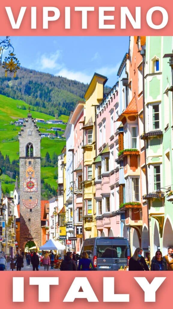A busy street scene in Vipiteno, Italy, features colorful buildings, a historic clock tower, and people engaging in daily activities against a backdrop of green hills, showcasing a destination in the Dolomites and a picturesque European village ideal for travel.